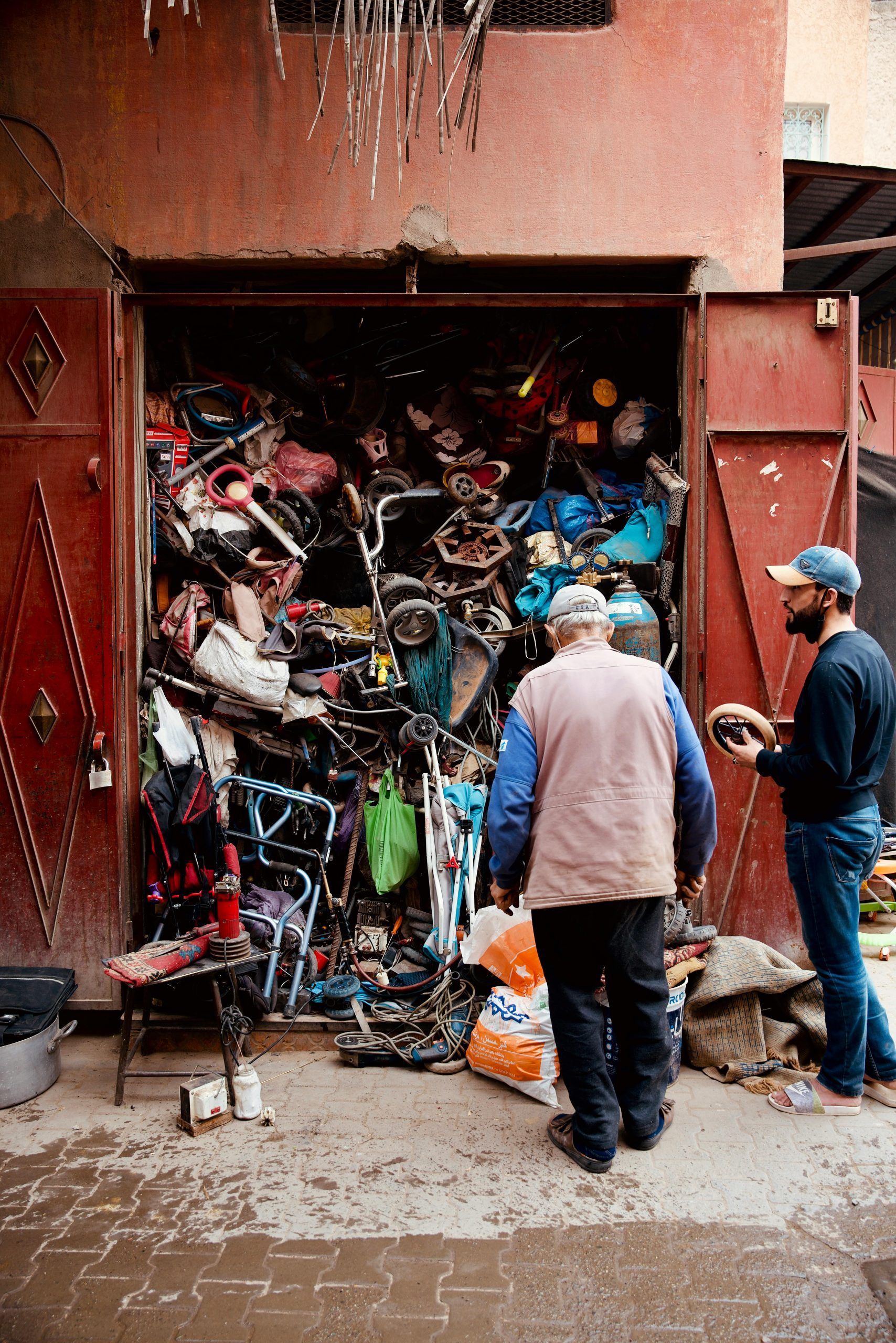 Two men inspecting a workshop with a cluttered collection of discarded items and tools.